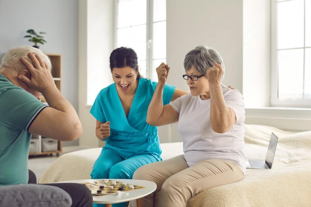 Happy senior patients playing checkers. Old retired couple and caretaker enjoy intellectual table games. Leisure activities as Alzheimer disease and dementia prevention therapy in