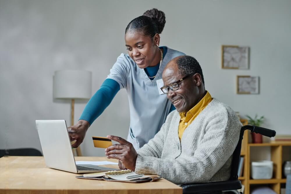 Young caregiver pointing at laptop and explaining man with disability how to pay online with credit card